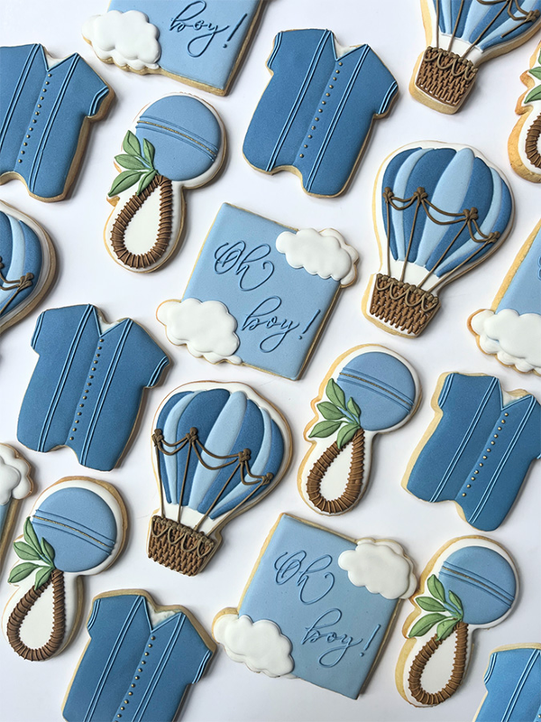 Decorative cookies shaped like baby onesies, hot air balloons, and rattles on a white background.